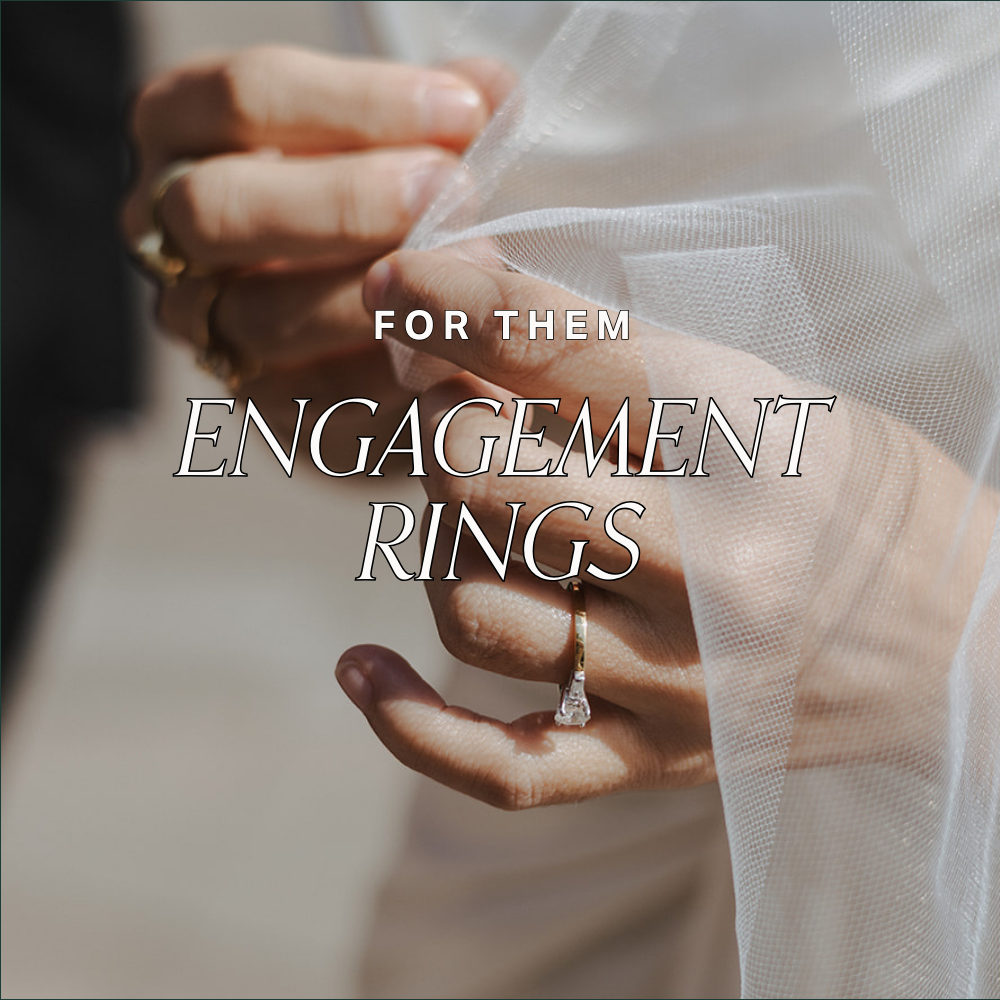 Close up image of hands weaved within a white veil with the focus on a vintage, yellow gold solitaire diamond ring with baguette diamond accents.Text reads: For them. Engagement Rings