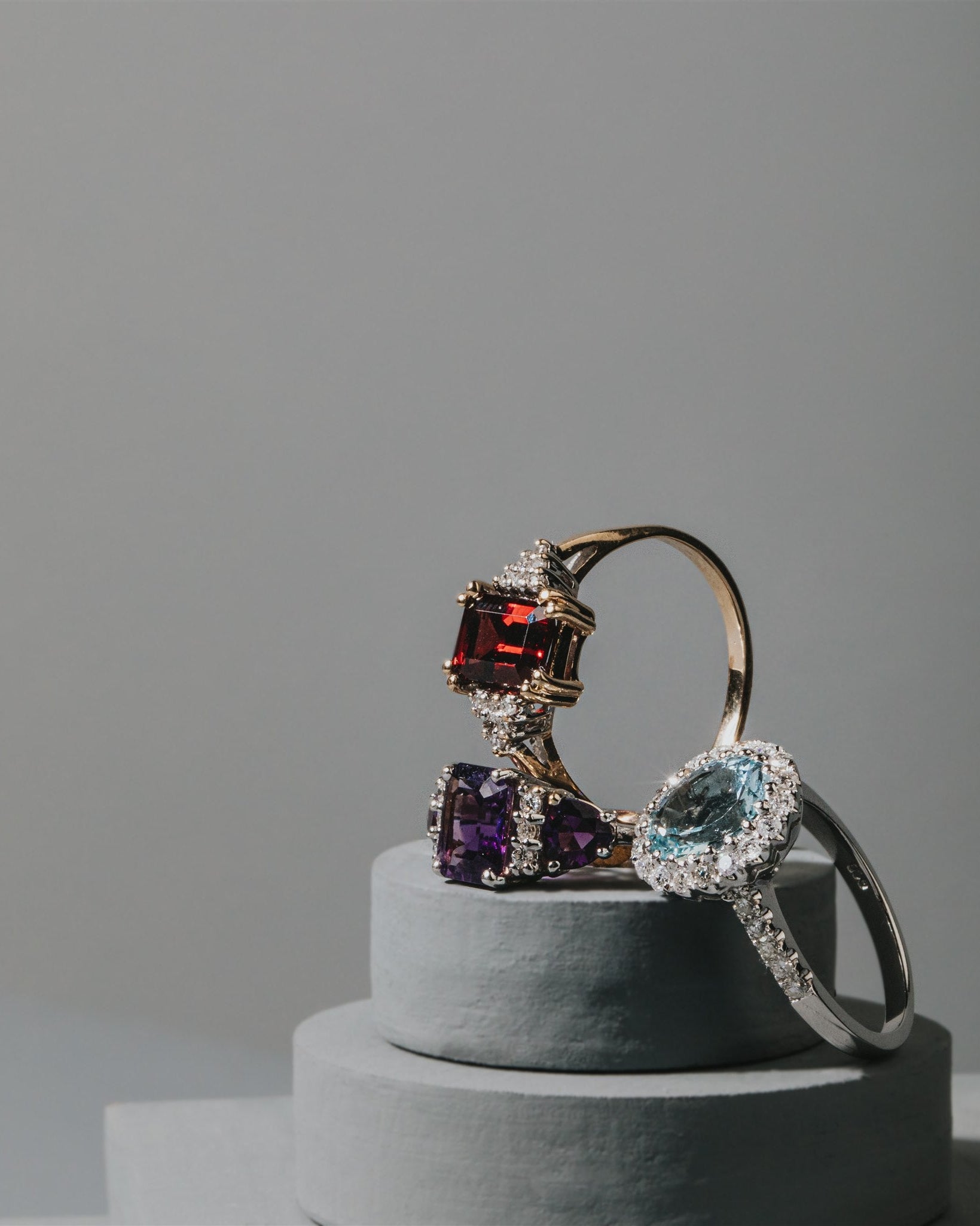 Three rings with colorful gemstones on a gray pedestal against a gray background