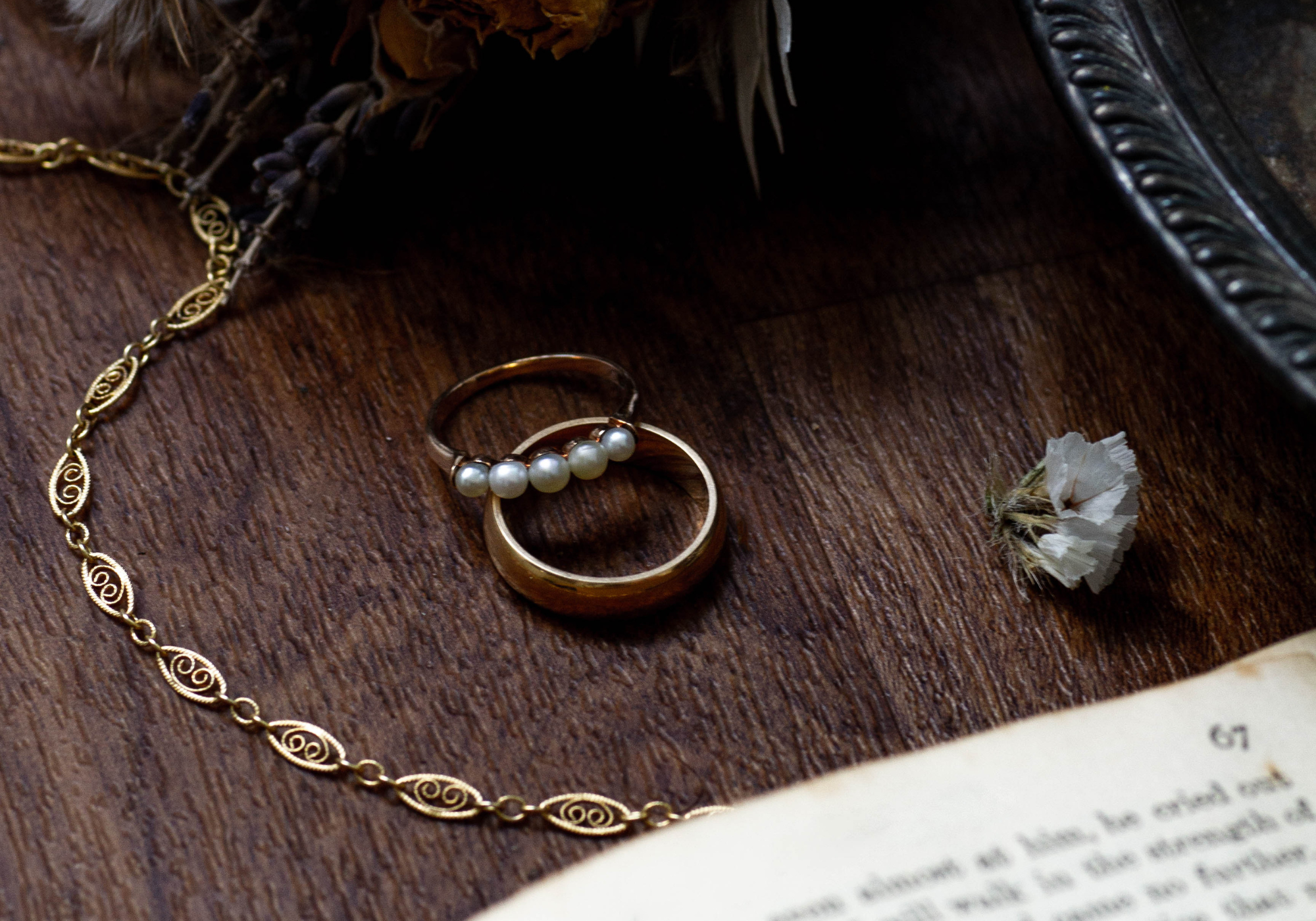 Flat lay of vintage gold rings, including a pearl-accented band, paired with an ornate gold chain necklace on a wooden surface, styled with dried flowers and an open book