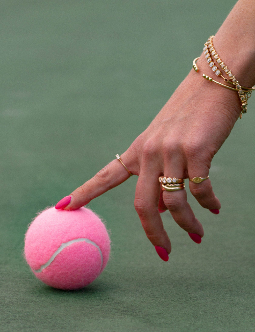 Image of hand touching pink tennis ball. Hand adorned in vintage, antique. yellow gold diamond, bracelet and ring stacks