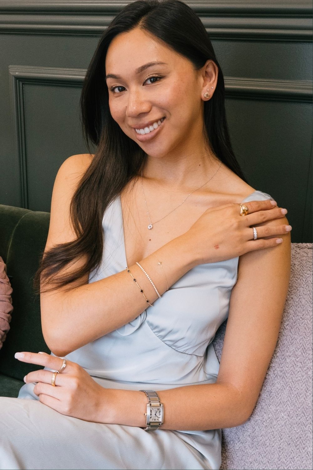A smiling woman with long dark hair sits on a green velvet sofa wearing multiple delicate, vintage jewellery pieces, including a two-tone Cartier watch, layered tennis bracelets, and stacked rings.