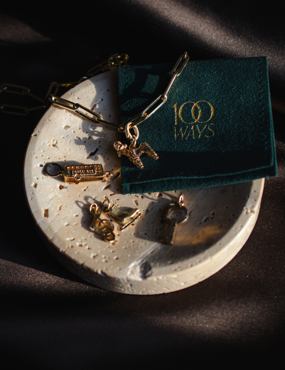 Vintage gold charm, paperclip chain necklace with miniature charms styled beside green velvet 100 Ways jewellery pouch on travertine dish