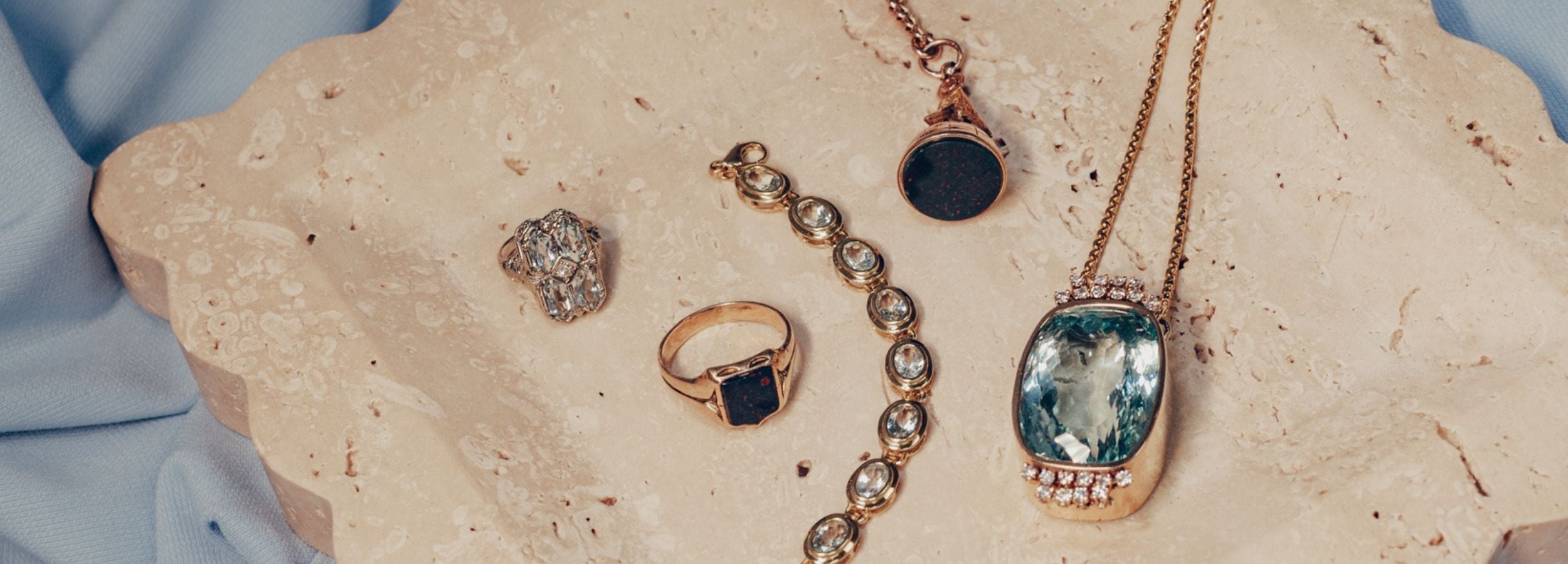 Vintage aquamarine jewellery styled with bloodstone signet ring and watch fob on travertine scalloped edge tray and light blue chiffon fabric