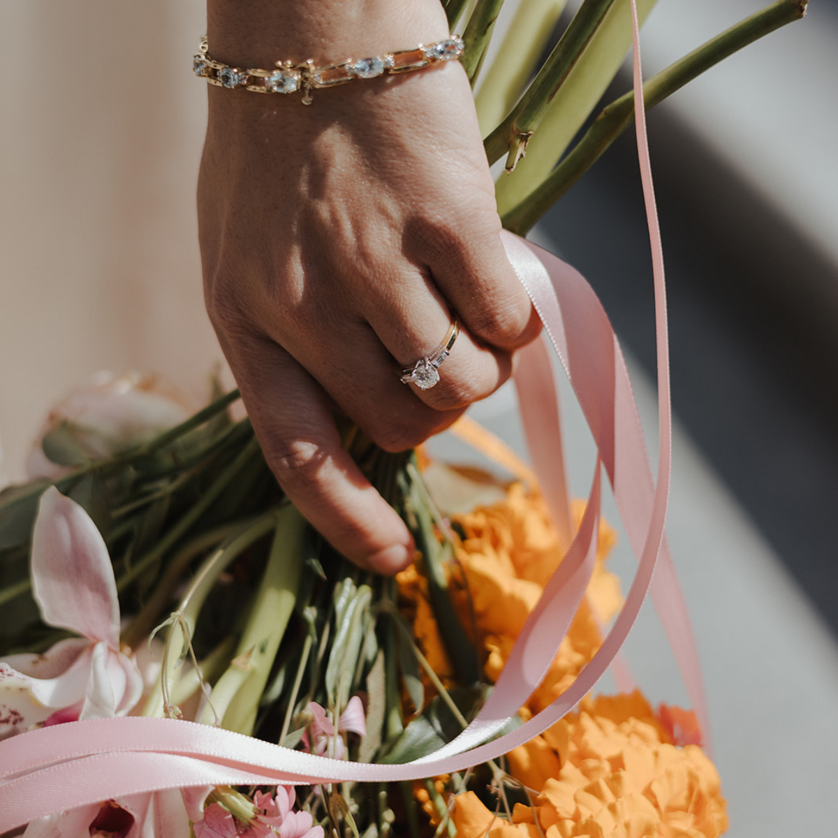 A close-up of a bride's hand holding a bouquet of vibrant marigolds and orchids, adorned with a vintage-inspired solitaire engagement ring and an elegant gold bracelet with aquamarine gemstones — both reflecting the charm and character of preloved, sustainable jewellery.
