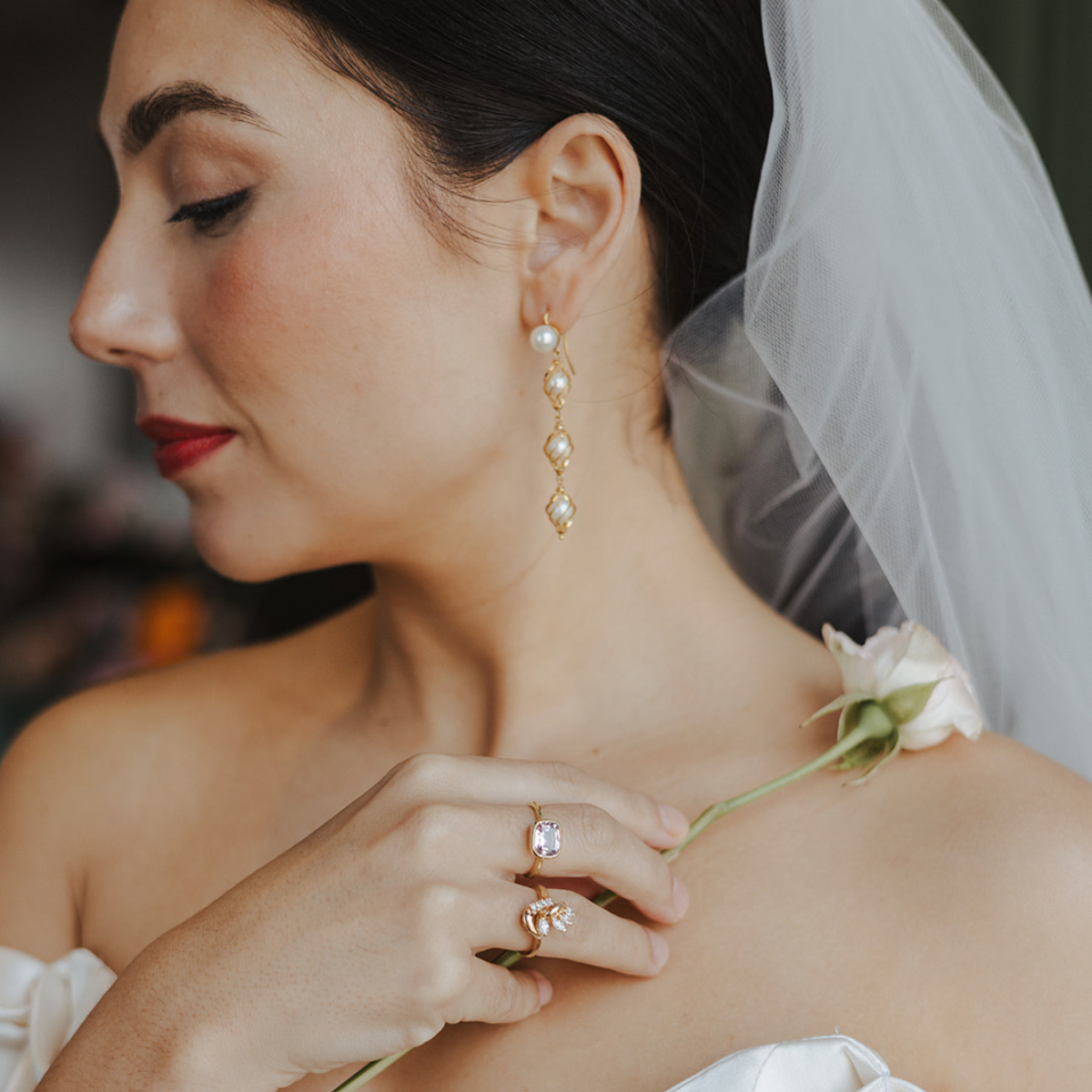 A bride wearing a soft off-shoulder gown and sheer veil, showcasing her vintage pearl drop earrings and statement rings. The ethically sourced, preloved jewellery pieces add timeless elegance and conscious luxury to her bridal look.