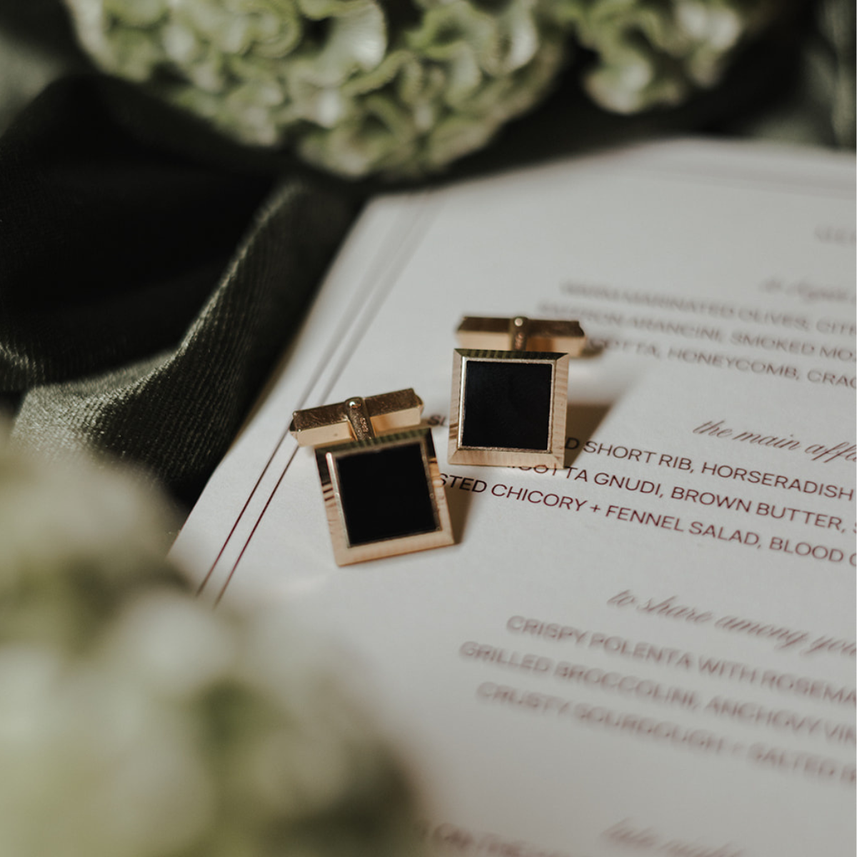 Yellow gold square cufflinks with onyx accents on a menu with greenery and olive green velvet in the background on top of a menu