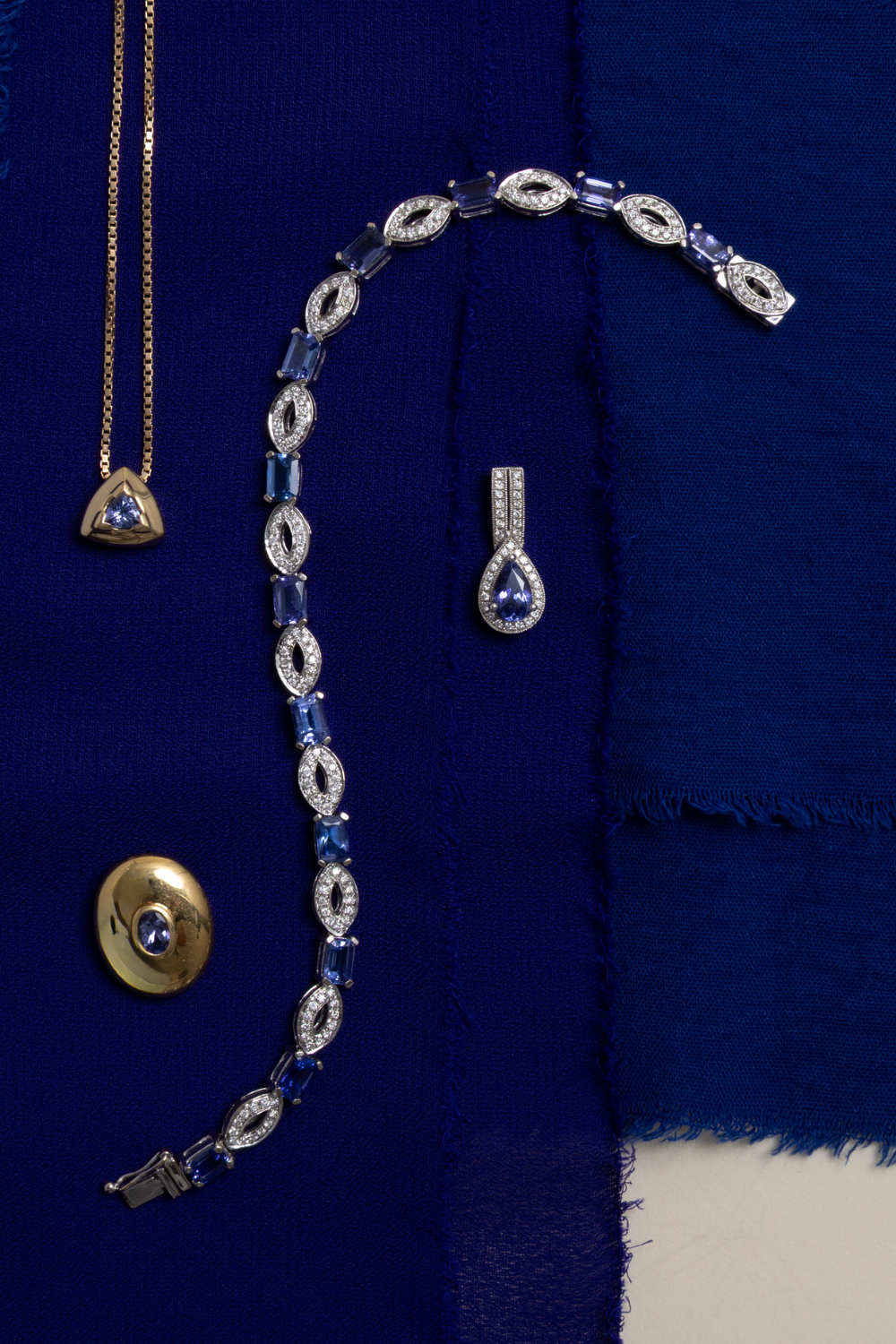 Preloved tanzanite and diamond fine jewellery flatlay featuring bracelet, necklace, and pendant on deep blue fabric.