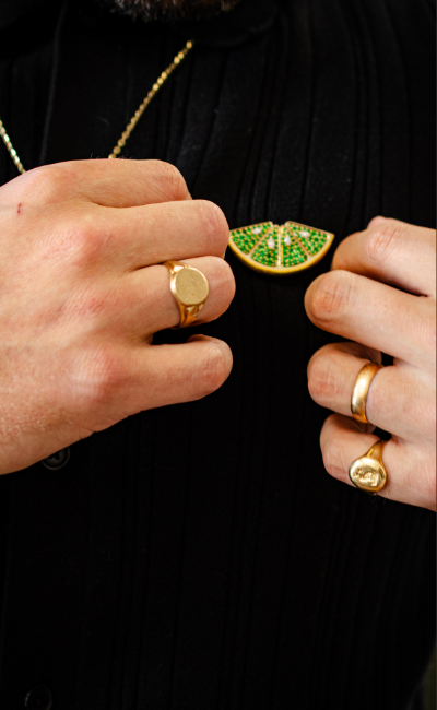 Close-up lifestyle shot of men's vintage jewelry, showcasing a solid gold classic signet ring, a thick yellow gold band, an engraved pinky ring, and a unique lime green wedge brooch pinned to a black ribbed shirt.