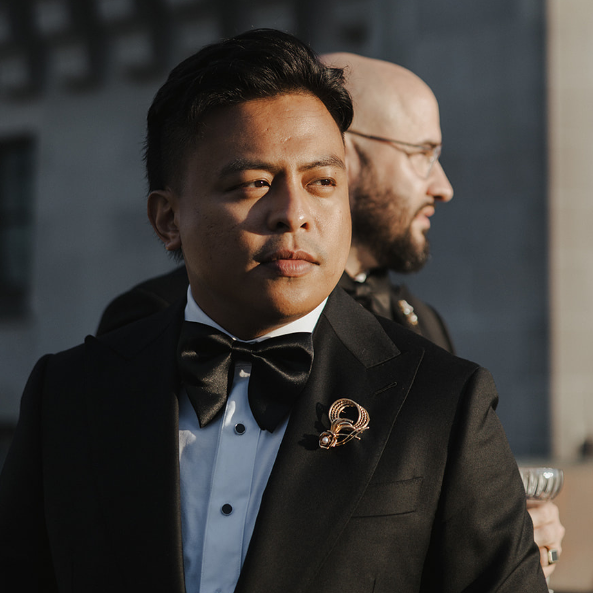 A groom in a classic black tuxedo, accented with two bold, vintage gold brooch on his lapel. The sustainably sourced, secondhand accessory adds a refined, personal touch to his formalwear, embodying elegance.