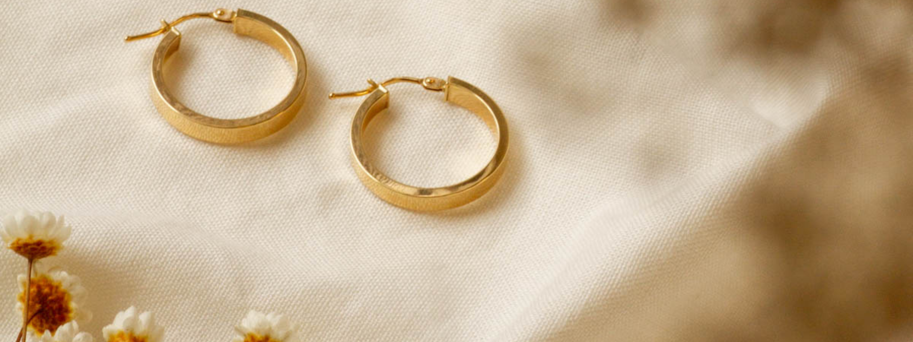 Yellow gold, minimal hoop earrings on a textured beige surface with blurred flowers in the background