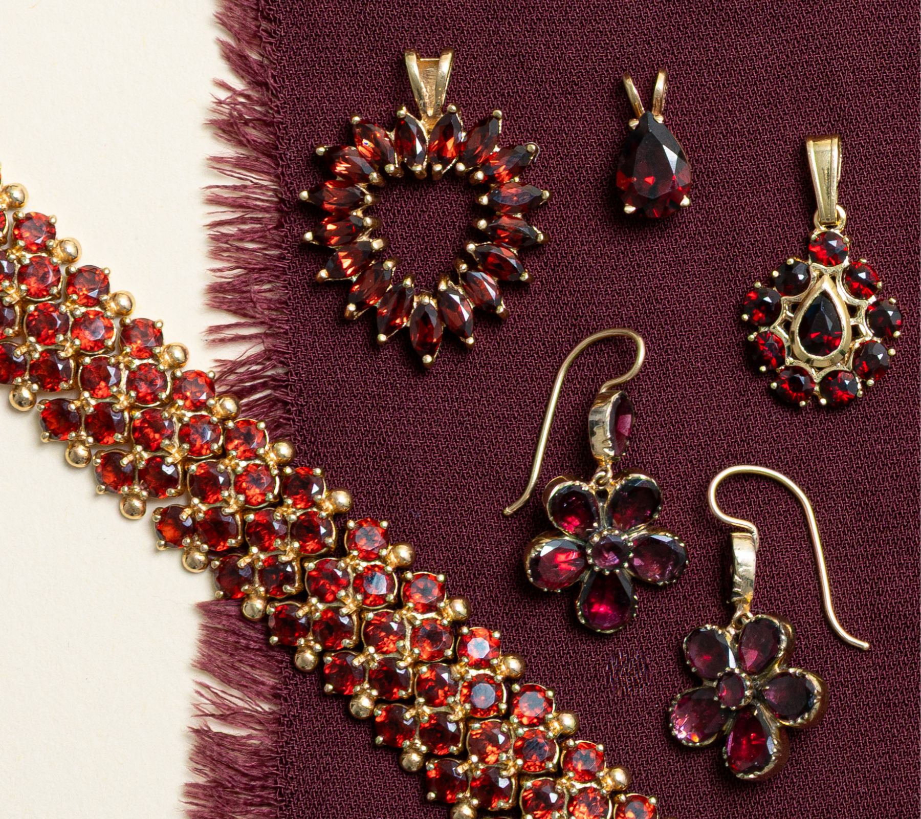 Vintage garnet jewellery laid on burgundy fabric, featuring chunky garnet gemstone bracelet, flower drop earrings and deep red pendants.