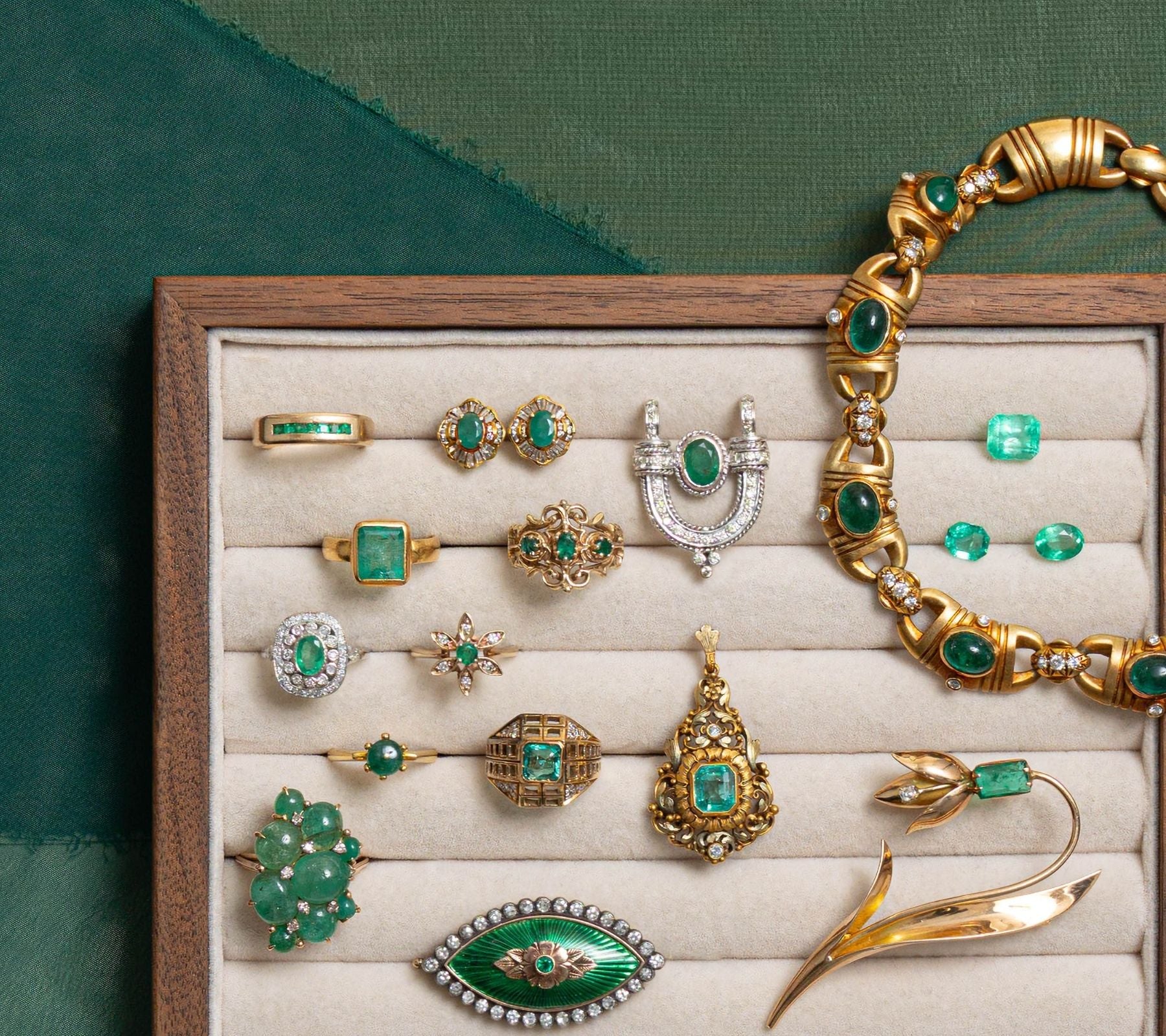 Curated collection of preloved emerald jewelry displayed on a walnut tray including rings, gold necklaces, vintage brooches, and loose emerald gemstones.