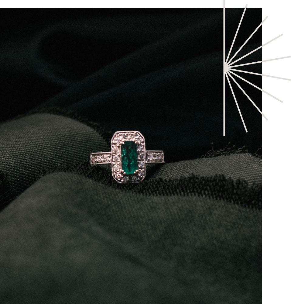 Antique, art deco inspired emerald and diamond cluster ring design showcased against a dark green fabric with a starburst graphic
