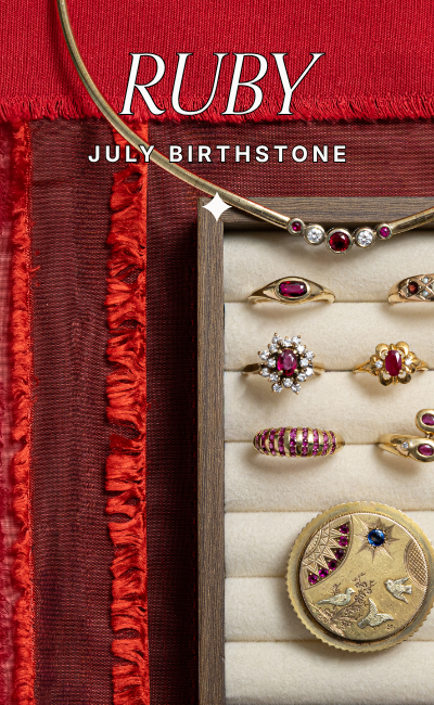 Curated selection of vintage ruby rings, a yellow gold collar necklace with ruby and diamond stones, and an antique engraved brooch with natural rubies in a velvet display tray. Text reads: Ruby, July Birthstone