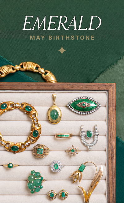 A wooden jewelry tray filled with vintage emerald and gold jewelry, including rings, a chunky bracelet, an unique shaped brooch, and a floral gold pin, set against a dark green chiffon. Text reads: Emerald May Birthstone.