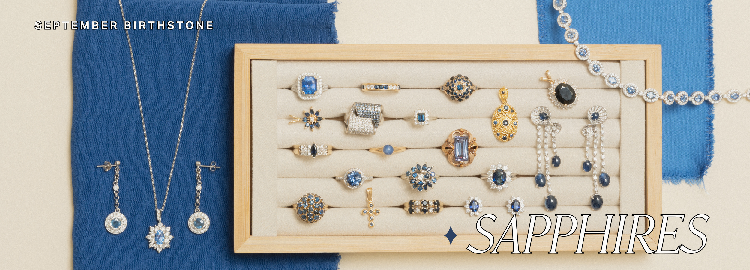 Display of vintage sapphire jewelry for September birthstone, featuring a wooden tray filled with ornate rings, brooches, earrings, and pendants adorned with blue sapphires and diamonds. Surrounding the tray are a sapphire pendant necklace, drop earrings, and a sapphire tennis bracelet, all set against rich blue and beige fabric backgrounds. A luxurious collection of antique gemstone jewelry highlighting intricate gold and silver settings with deep blue sapphires.