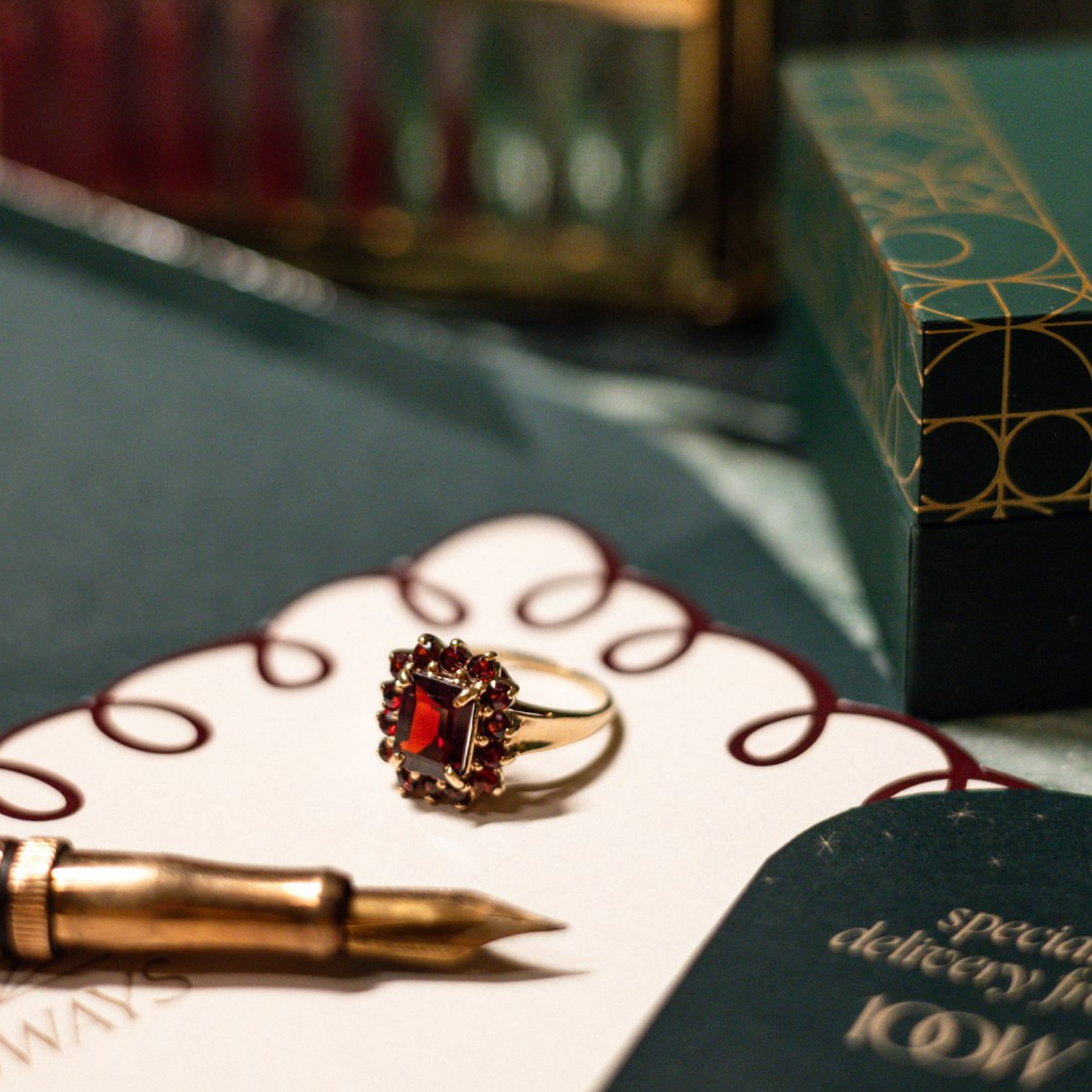 Vintage gold ring with deep red, garnet gemstone and halo setting, styled beside a fountain pen and green 100 Ways jewellery box
