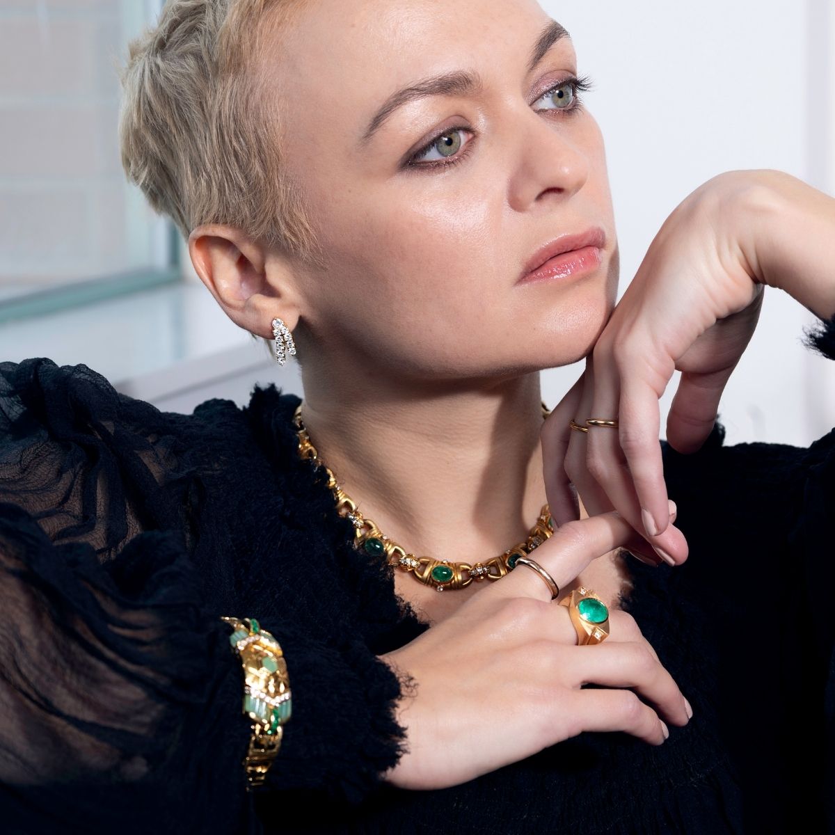 Model wearing a black top styled with a chunky gold and emerald link necklace, emerald cocktail ring, and diamond earrings.