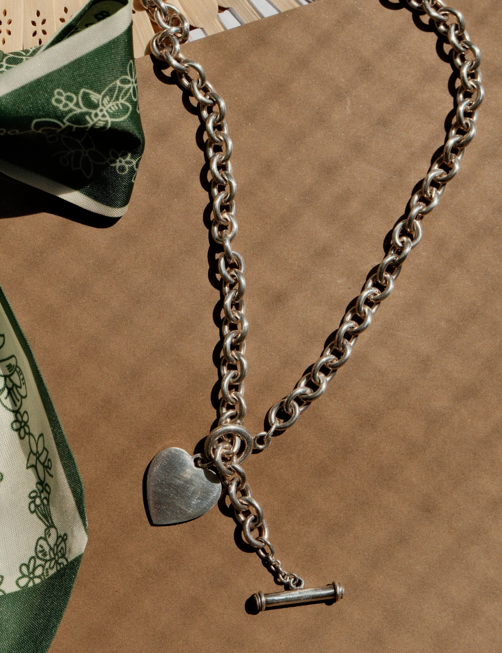 Chunky vintage sterling silver chain necklace featuring a large heart pendant and toggle clasp, laid next to a green patterned silk scarf on a natural brown background.