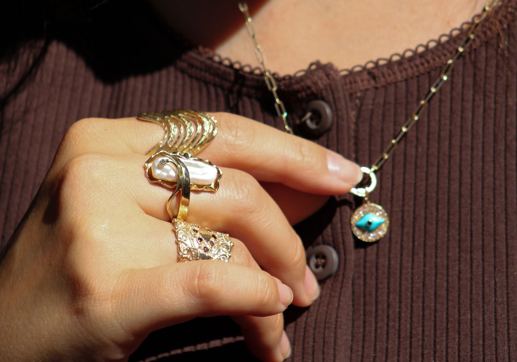 Close-up of a hand resting on a brown ribbed shirt, wearing three vintage yellow gold statement rings, including one with a baroque pearl, and is touching a gold necklace featuring a diamond-studded evil eye pendant with a turquoise center.
