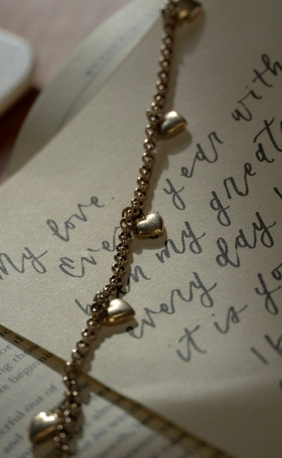 Delicate gold heart charm bracelet draped across a handwritten love letter, romantic vintage jewellery detail with soft, warm lighting.