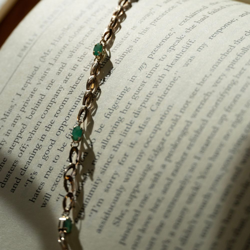 Vintage gold link bracelet with oval emerald gemstones draped across an open book page, romantic antique jewellery styled with warm, moody lighting.