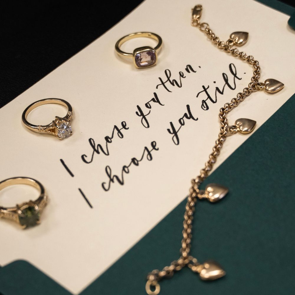 Vintage-inspired gemstone and diamond rings arranged on a handwritten love letter card beside a gold heart charm bracelet, romantic heirloom fine jewellery.