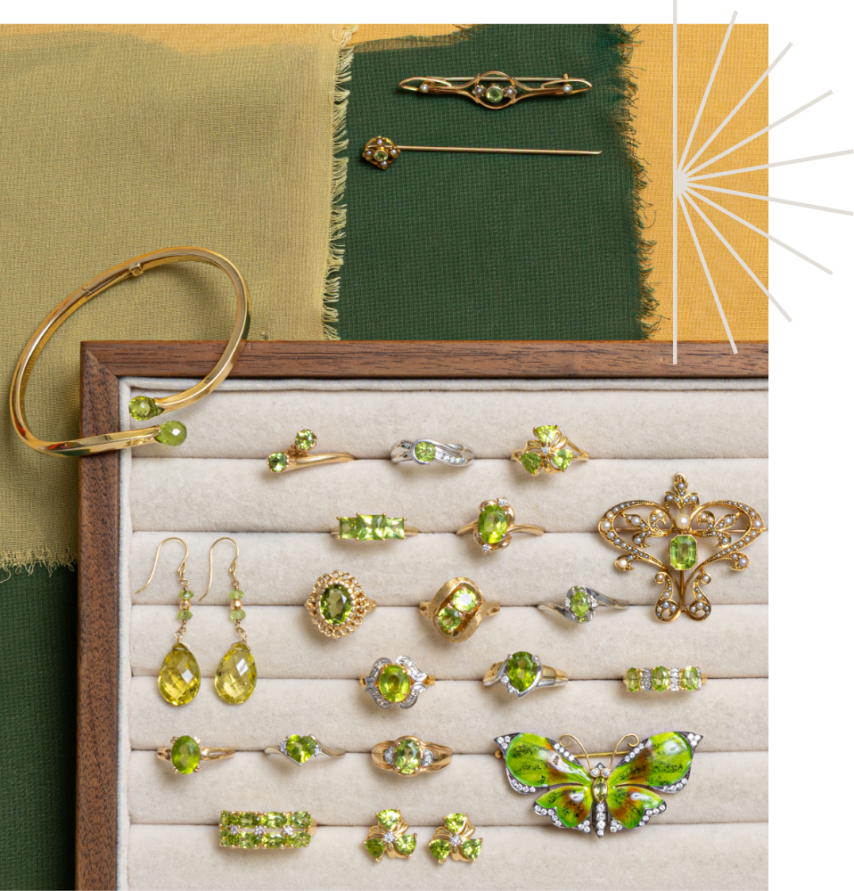 A jewelry display tray filled with vintage and antique peridot rings, earrings, brooches, and a butterfly brooch. A gold bangle and two brooches are placed above the tray on fabric swatches.