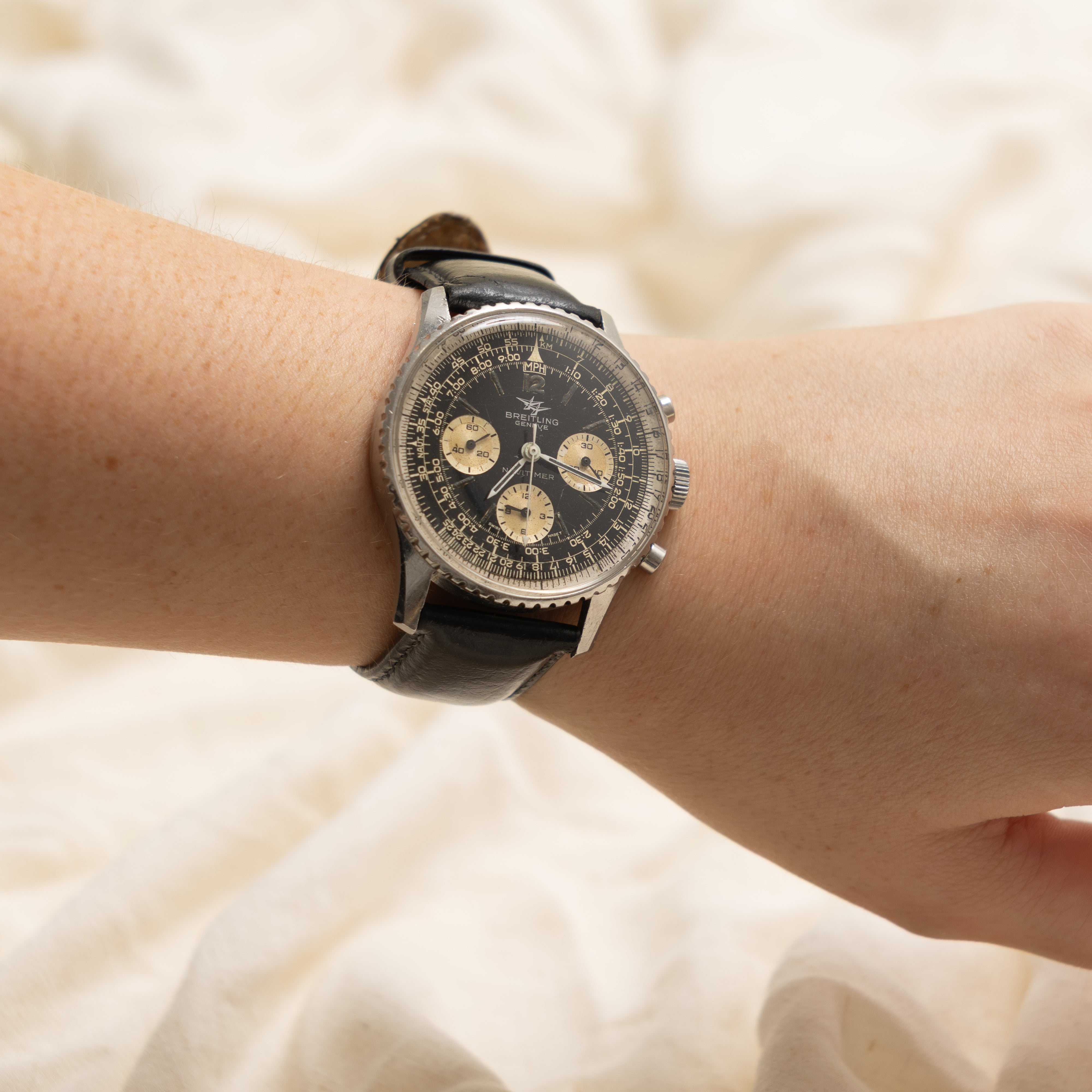 A vintage-style wristwatch with a black dial, three subdials, and a black leather strap, shown worn on a person's wrist against a soft cream fabric background.