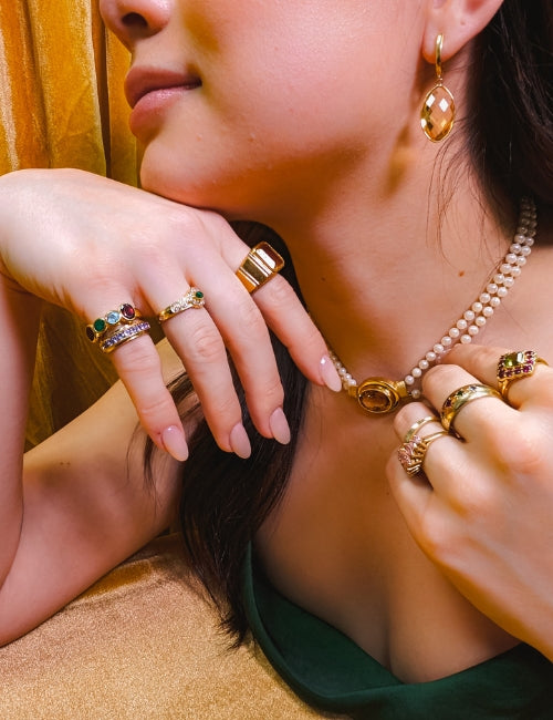 image of woman in front of golden yellow backdrop. woman is wearing several bold gemstone rings and other fine jewellery pieces