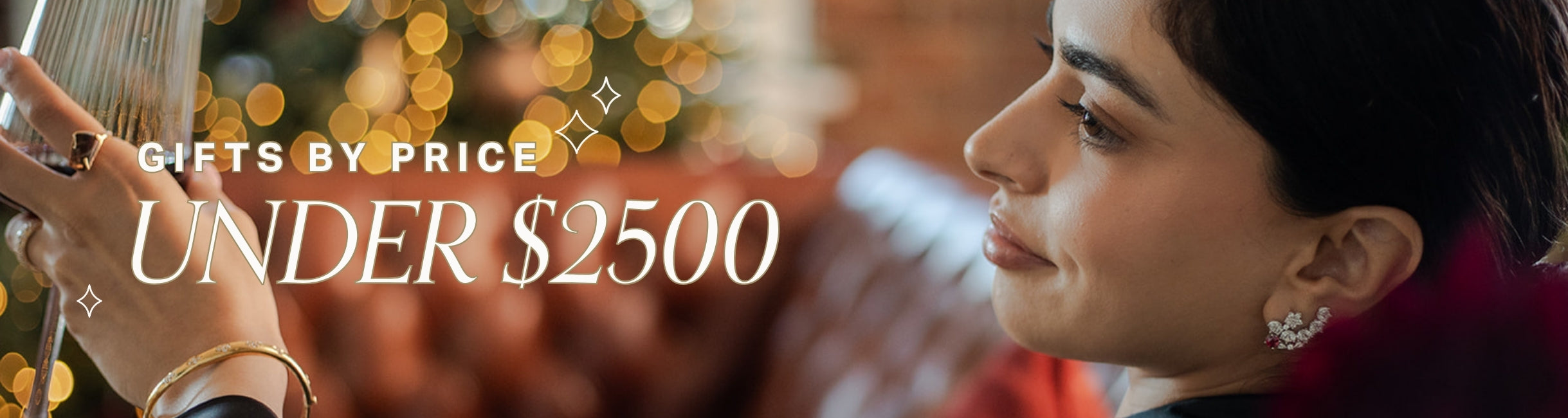 Woman holding a wine glass, wearing gold and gemstone jewellery with blurred holiday lights and text overlay: gifts by price, under $2500