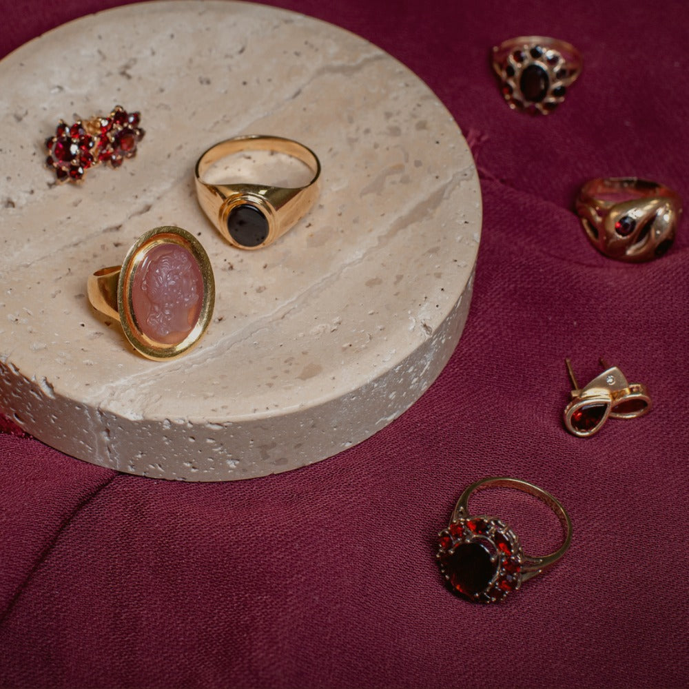 Jewellery set including ruby red and carnelian rings and earrings on a round stone surface with a maroon fabric background.
