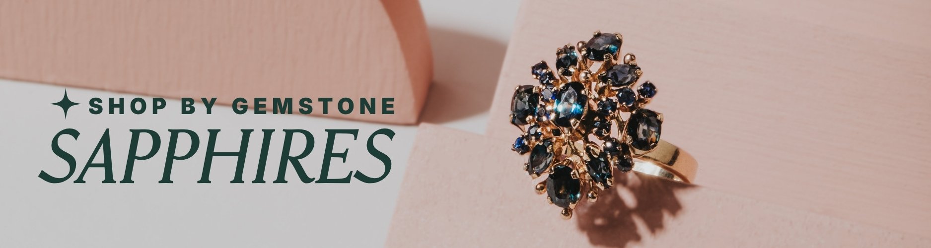 Gold ring with sapphire stones on a pink background, text overlay reads: 'Shop by Gemstone, Sapphires'