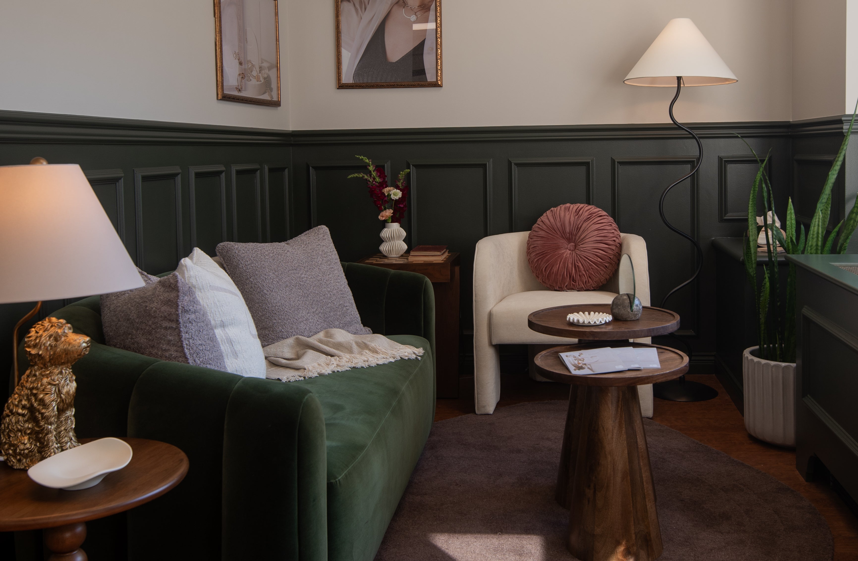 A cozy showroom for a vintage, preloved jewelry brand featuring a green velvet sofa, cream accent chair, wooden tables with decor, and elegant touches like floral arrangements, framed portraits, and warm lighting in a refined, vintage-inspired setting.