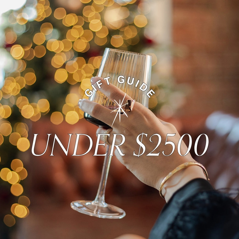 Woman holding a wine glass, wearing gold and gemstone jewellery with blurred holiday lights and text overlay: gifts by price, under $2500