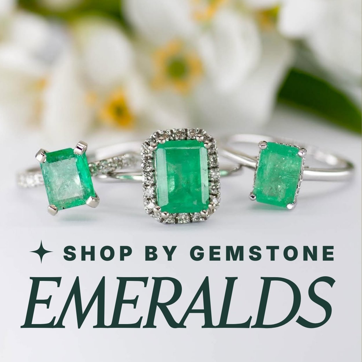 Three emerald rings in white gold settings on a white background with out of focus florals in background. Text overlay reads: 'Shop by Gemstone: Emeralds'
