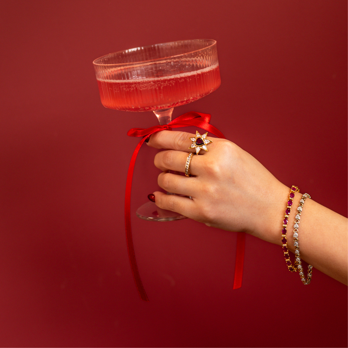 Red background with hand adorned with vintage, antique ruby and heart themed jewelry. Holding coupe glass with red bow and pink sparkling