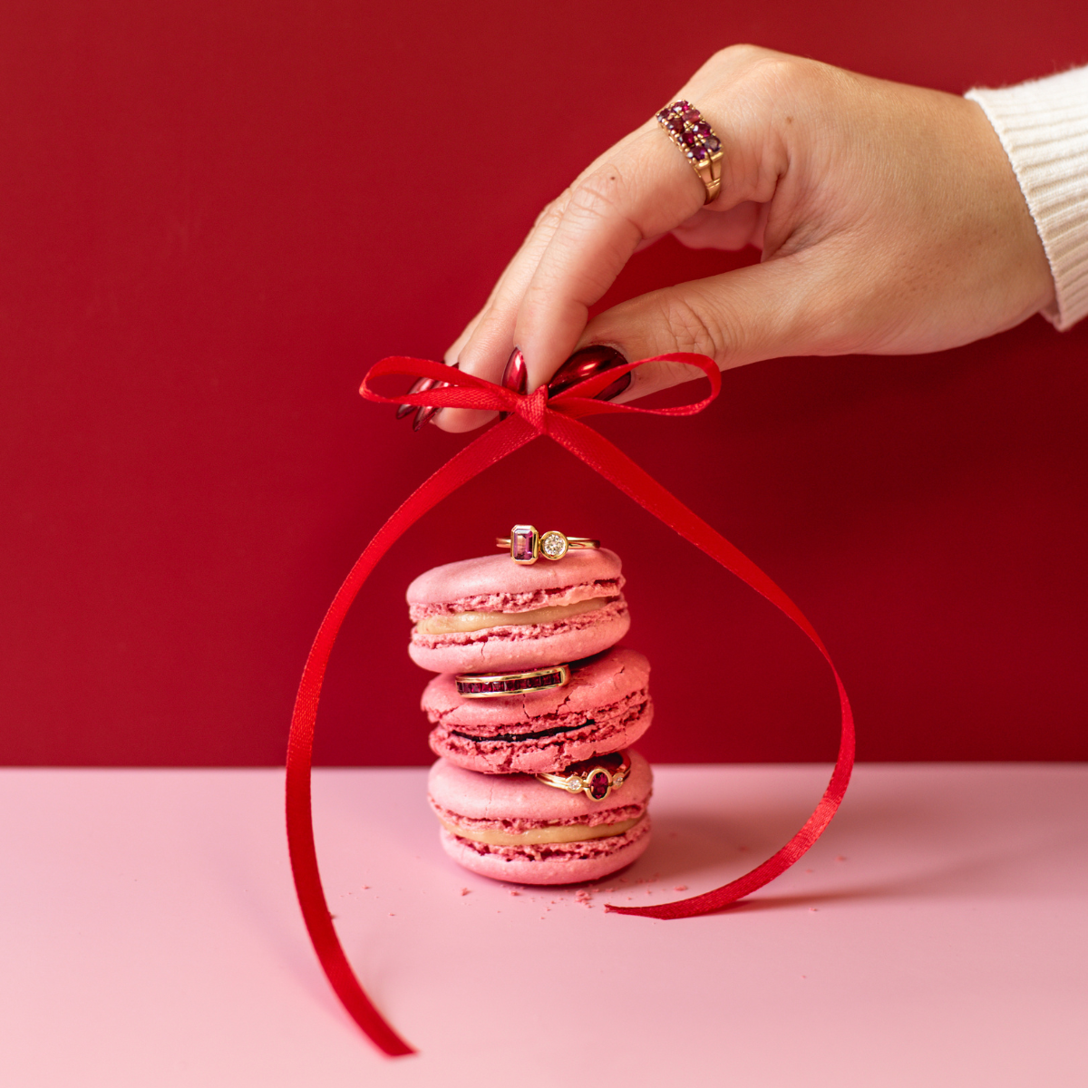 Stack of multi coloured pink macarons with ruby vintage rings inbetween layers and hand holding red satin ribbon on top