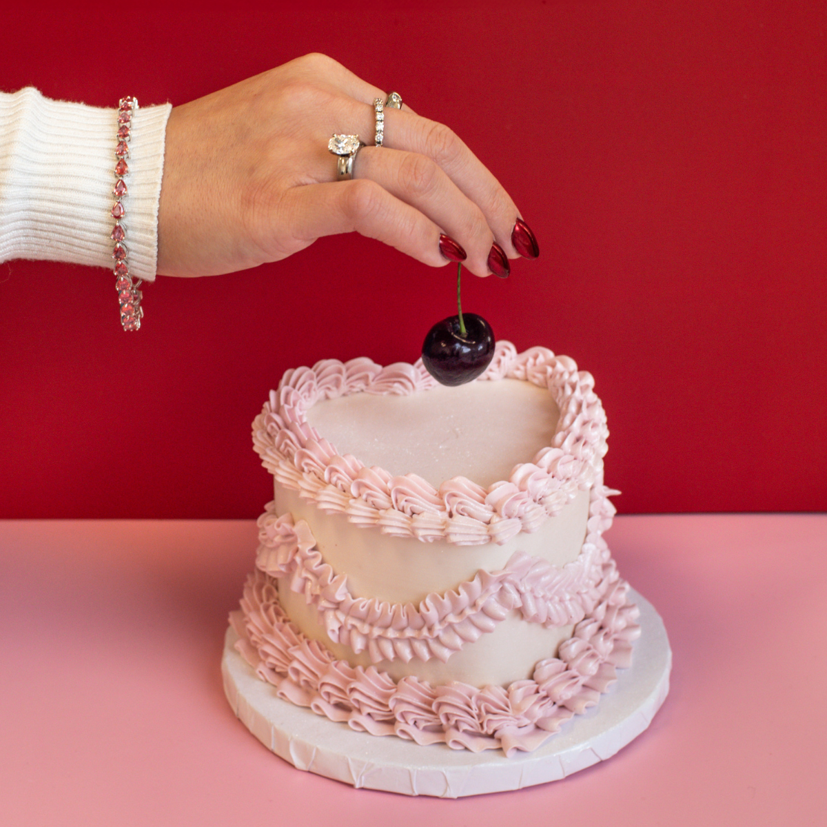 Vintage inspired mini heart cake with hand holding cherry on top. Hand model wearing preloved, antique white gold diamond jewelry and pink coloured sapphire tennis bracelet