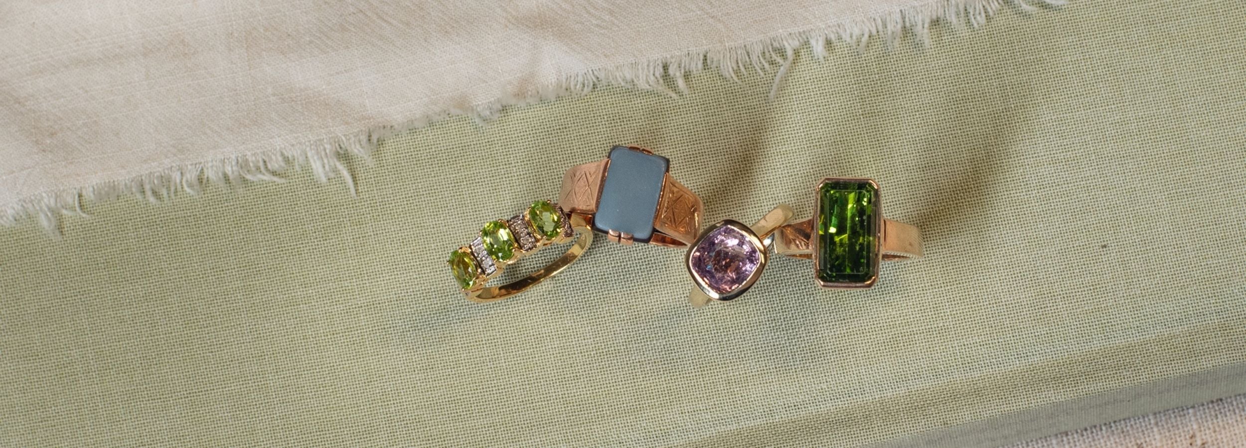 Three yellow gold rings featuring gemstones as August birthstones: peridot, sardonyx and spinel. Arranged on a light sage green textured fabric background.