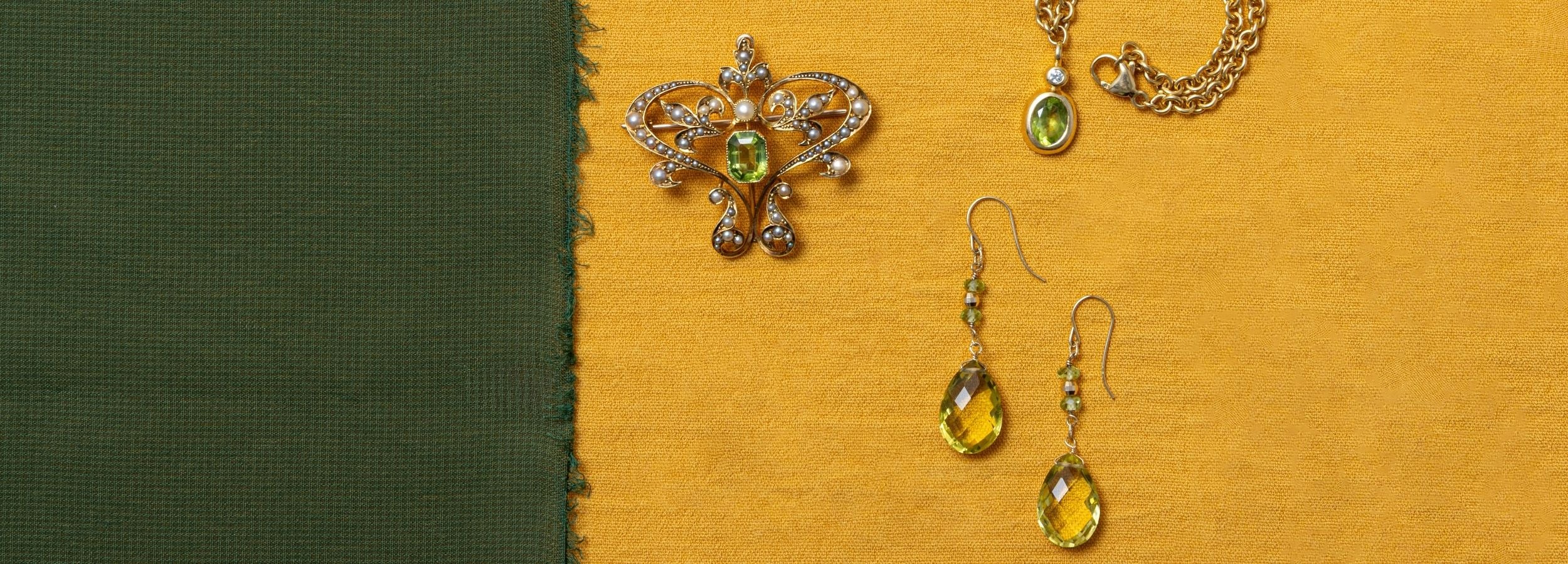 Jewellery flatlay featuring a vintage gold-tone brooch with green peridot gemstones, peridot drop earrings, and a yellow gold chain with a peridot gemstone at the end, arranged on a textured mustard yellow background with a dark green vertical panel on the left.
