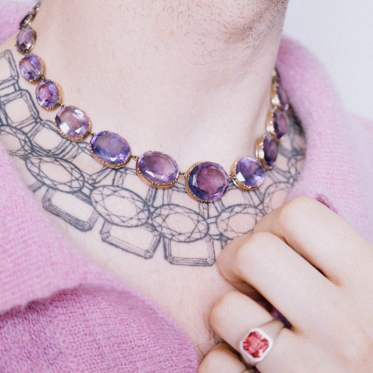 Detail shot of model wearing a chunky vintage, amethyst statement necklace, styled over a jewellery collar tattoo and pink sweater.