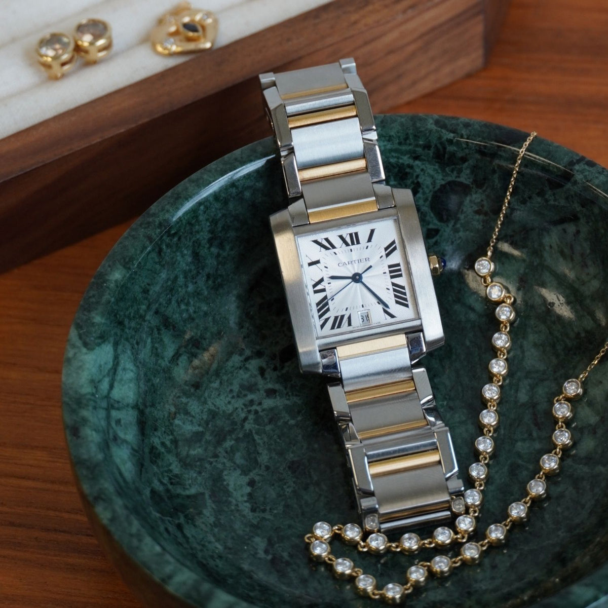 A Cartier Tank watch with a silver and gold-tone two-tone metal bracelet and a white dial, displayed on a green marbled bowl next to a yellow gold diamond tennis necklace. 
