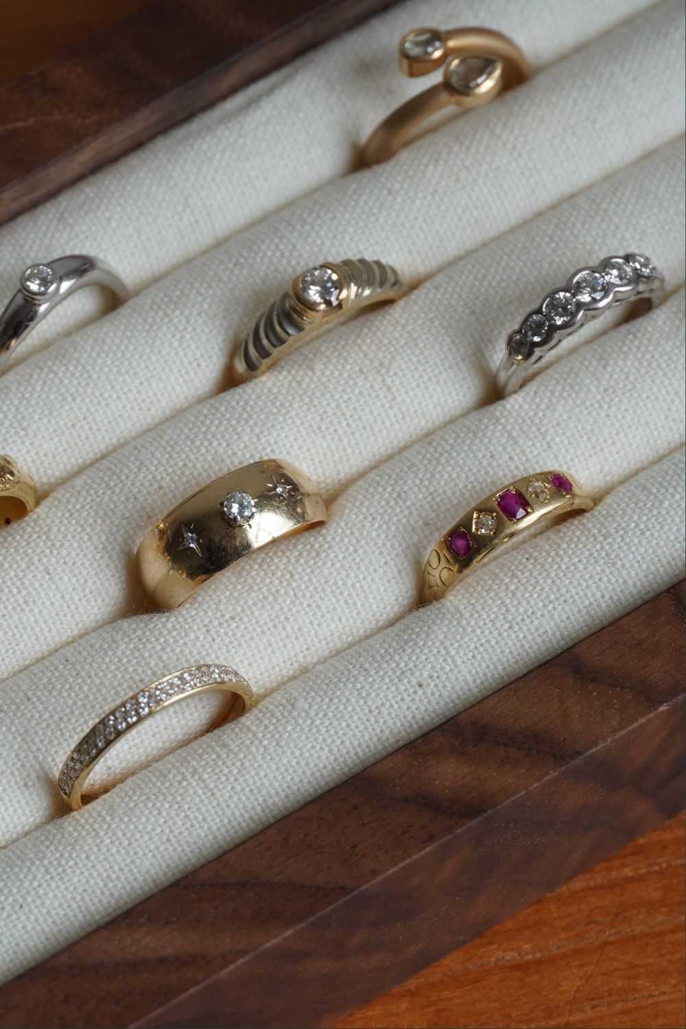 A close-up of multiple rings displayed in a cream fabric-lined jewelry tray. The vintage rings include yellow gold and white gold diamond bands, some with clear gemstones and one with 1883 Chester Five Stone Ruby & Diamond Ring.