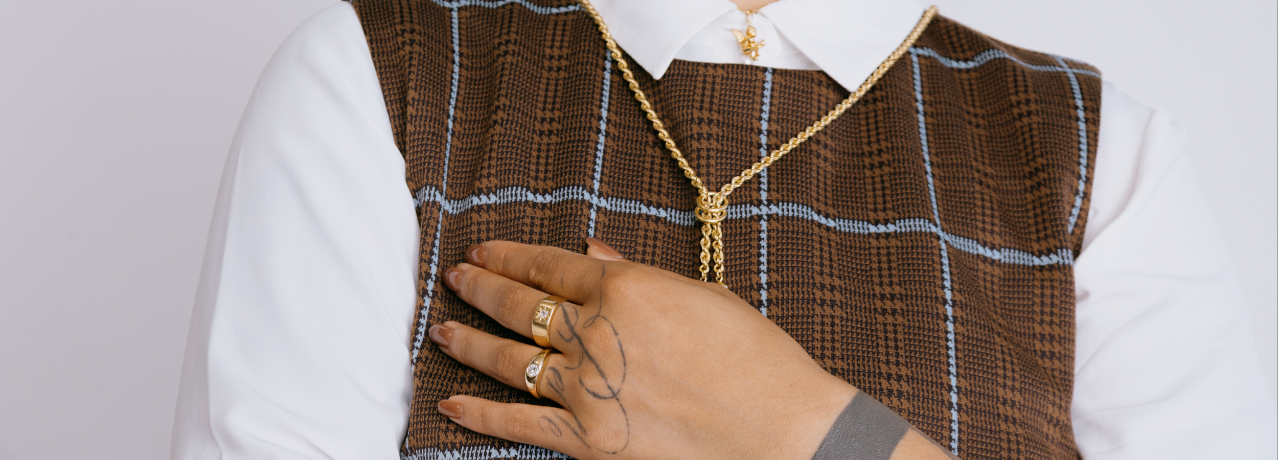 Close-up of a woman's hand resting on a brown plaid vest, showcasing two preloved vintage gold rings. One ring features a starburst setting and the other a dome diamond ring, paired with an antique, bolo style gold chain necklace.