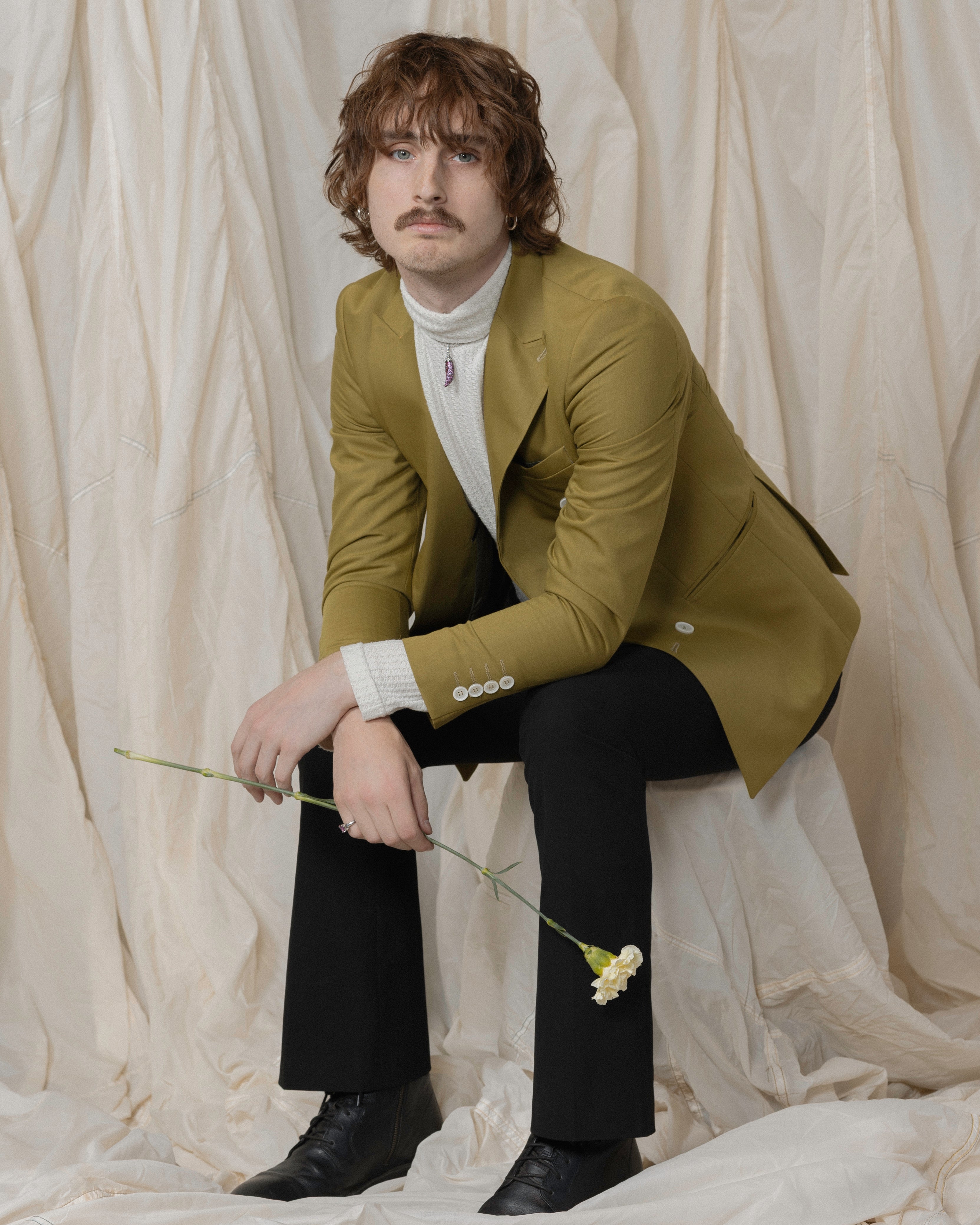 Model wearing olive blazer over a white turtleneck and black trousers, holding a single white carnation. Wearing subtle, vintage jewelry, including hoop earrings and chilli pendant necklace. Shot against soft neutral draping for the Vancouver Pride 2025 jewelry collection. 