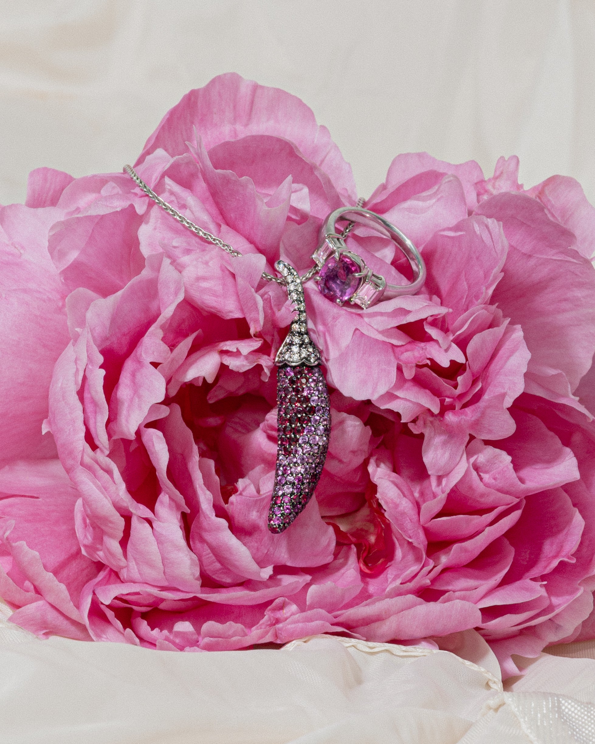 Close-up of a vivid pink peony flower styled with a sparkling pavé crystal chili pepper pendant and pink sapphire cocktail ring.