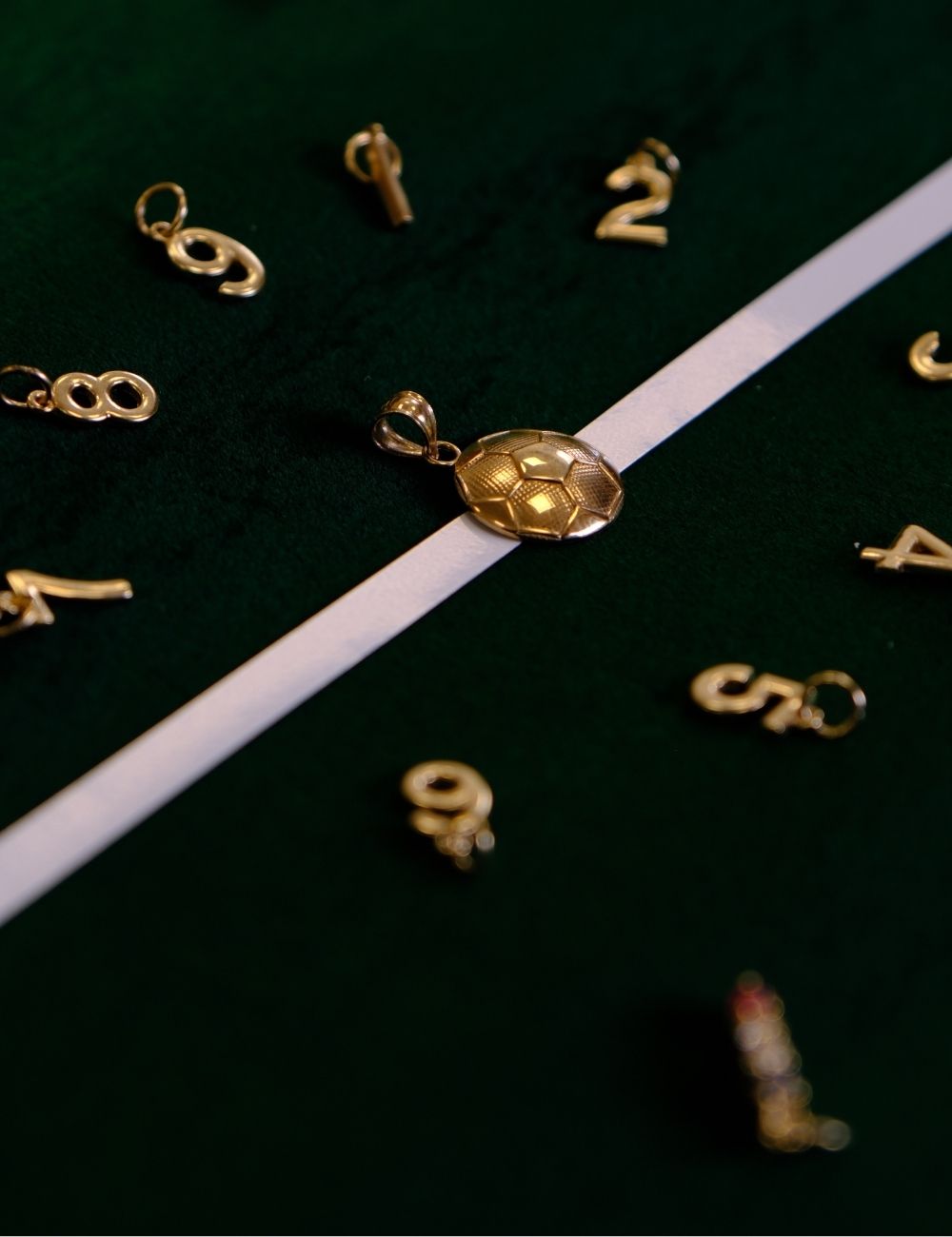 Gold soccer ball pendant and jersey number charms displayed on dark green velvet with white field line for a soccer inspired  jewelry collection.