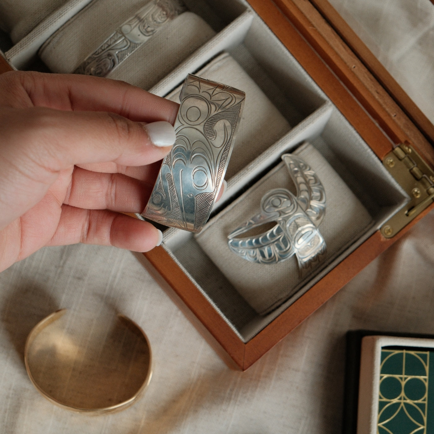 Holding a preloved vintage, indigenous silver cuff bracelet featuring engraved Indigenous art, displayed above a wooden jewelry box containing other indigenous jewellery.