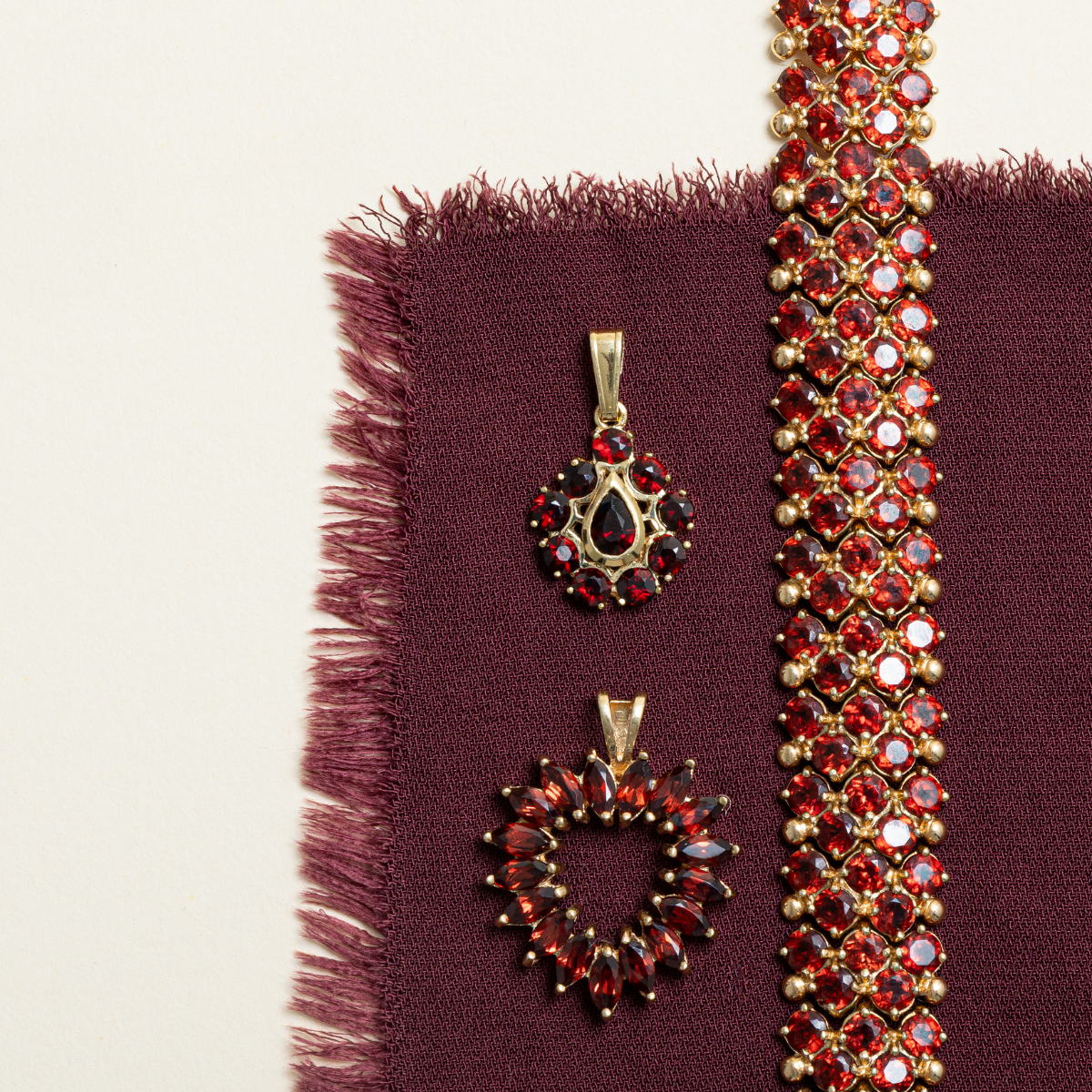 Vintage garnet pendants and bracelet in deep crimson tones, set in yellow gold and styled on textured burgundy fabric for January birthstone.